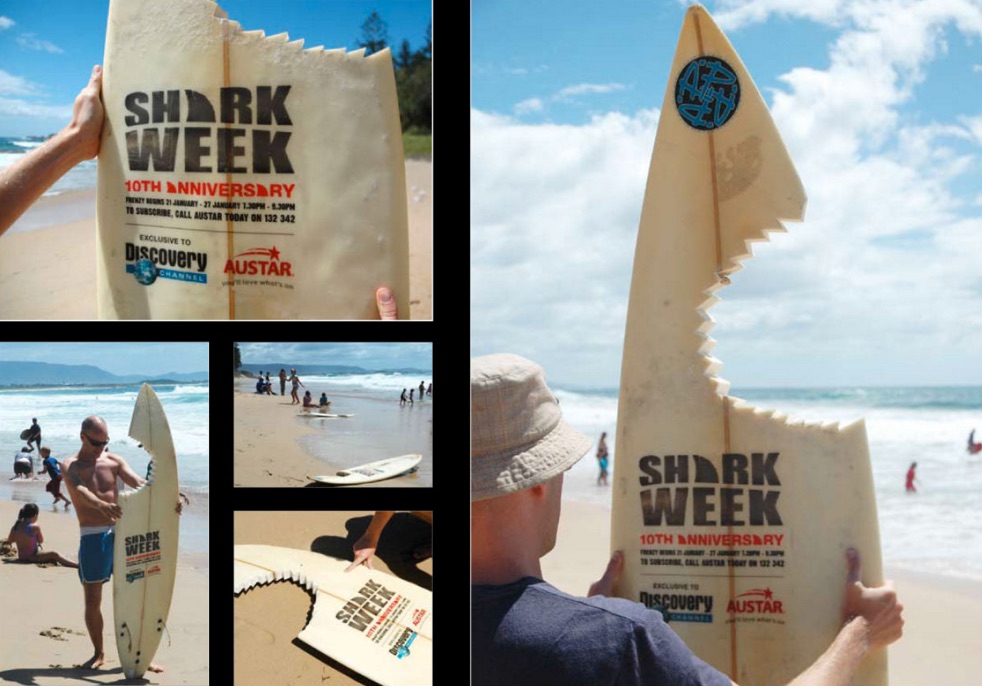 Pictures of a surfboard on a beach with a jagged chunk ripped out so that it looks like a shark chomped on it. The surfboard says Shark Week 10th Anniversary and has more details about the event.