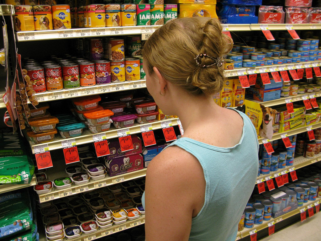 View of woman from behind, standing in front of grocery store shelves looking at dog food selection.