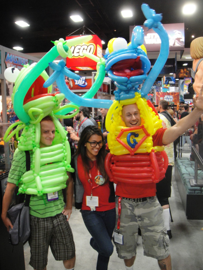 Two young guys pose on either side of a young woman. The guys are wearing colorful Kermit the Frog and Gonza costumes constructed out of balloons. The three are attending the San Diego Comic Convention.