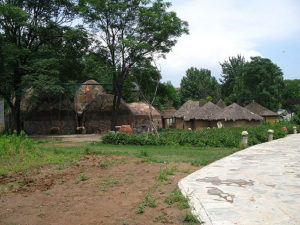 a cluster of thatched-roof circular buildings under trees, with a small garden in front of them