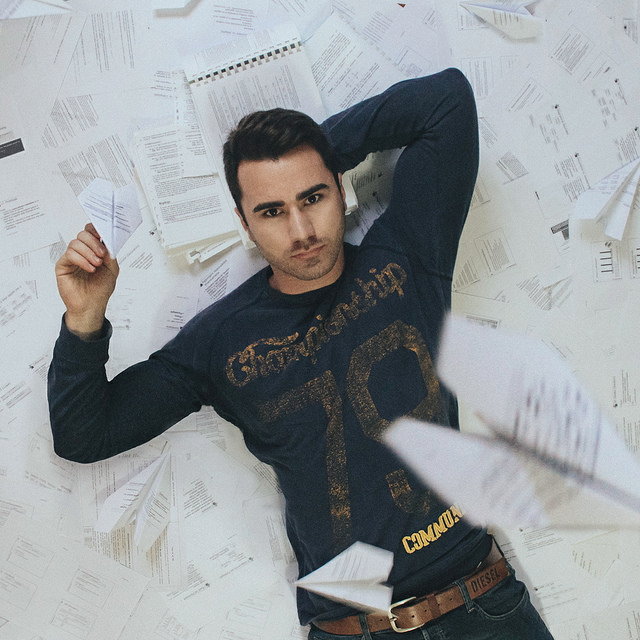 Young man laying on back in pile of papers, with a folded paper airplane in hand. Two additional paper airplanes are in air above him.