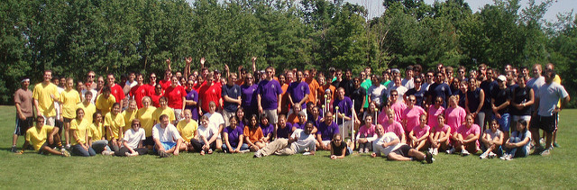 A large group of people posing for the camera outdoors. Clusters of people wear similarly colored shirts in yellow, red, purple, pink, and black