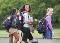 Two young women wearing backpacks walking outside among other students