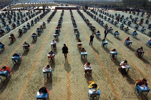 rows of students in desks outdoors, taking an exam. A couple of teachers walk between rows.