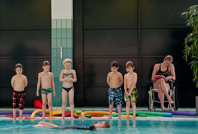 five children in swimsuits on the edge of a pool, watching an adult demonstrating a swim technique. A woman is seated with a paddleboard next to the children.