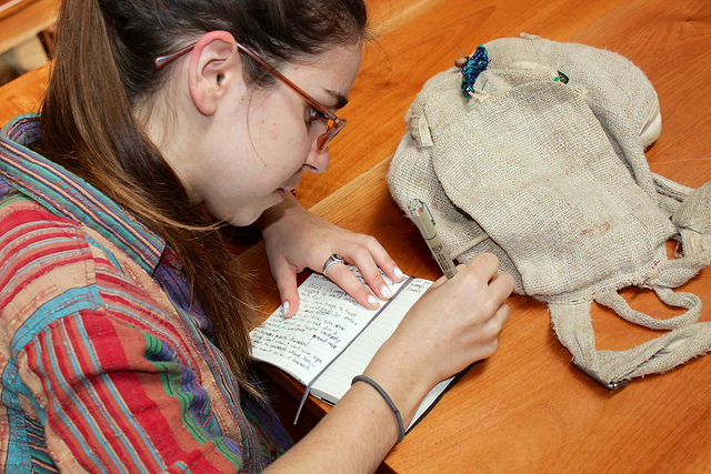 Photo looking over shoulder of young woman writing in a small notebook on a table