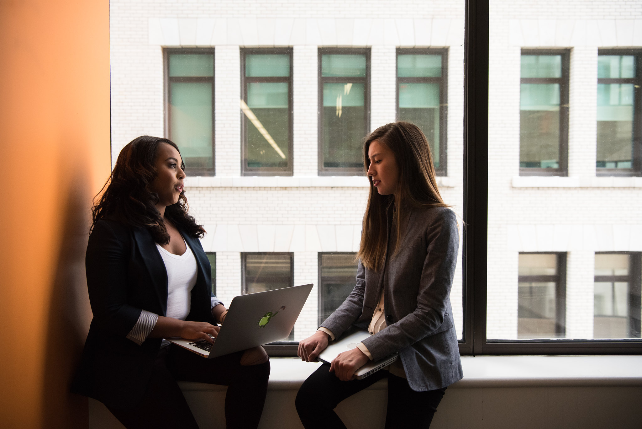 Photograph of two women sitting on a window bench. One is holding an open computer and talking to the other.