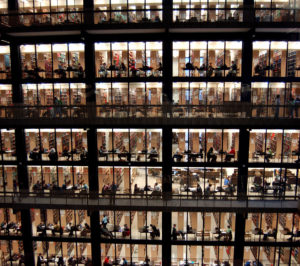 Sideview photo of eight floors of an office building, showing many people inside at their desks. Title of photo: Worker Bees.