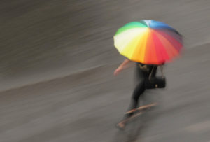 Photo of a woman running in the rain holding a rainbow-colored umbrella.