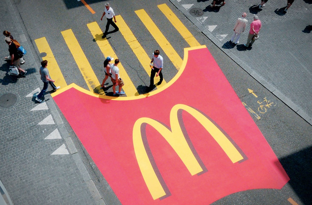 A McDonald's fries container is painted on the street near the yellow stripes of a crosswalk so that the crosswalk stripes appear to be fries.