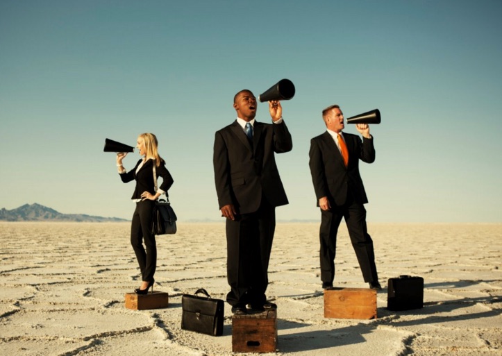 Three businesspeople stand in the middle of the desert, their briefcases at their feet. Each businessperson is shouting into a megaphone in different directions.