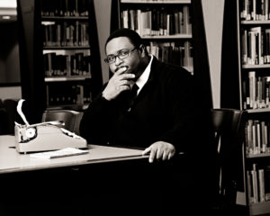 Photo of a man sitting in an office or library with a small typewriter on the table. His head rests on his hand, and he looks contemplative.