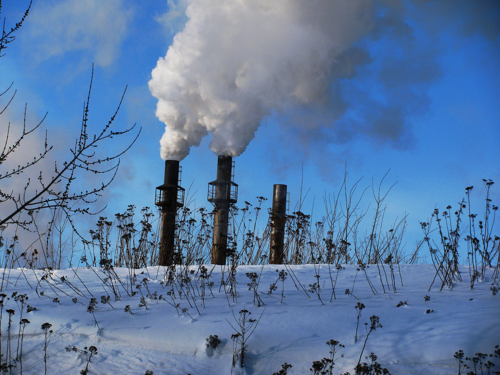 Photo of snowy landscape with three factory chimneys on the horizon, filling the air with smoke.