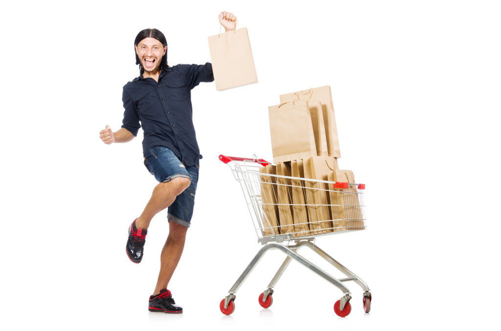 Photograph of a person excitedly shopping. Their shopping cart is full of bags, and they are holding one in the air.