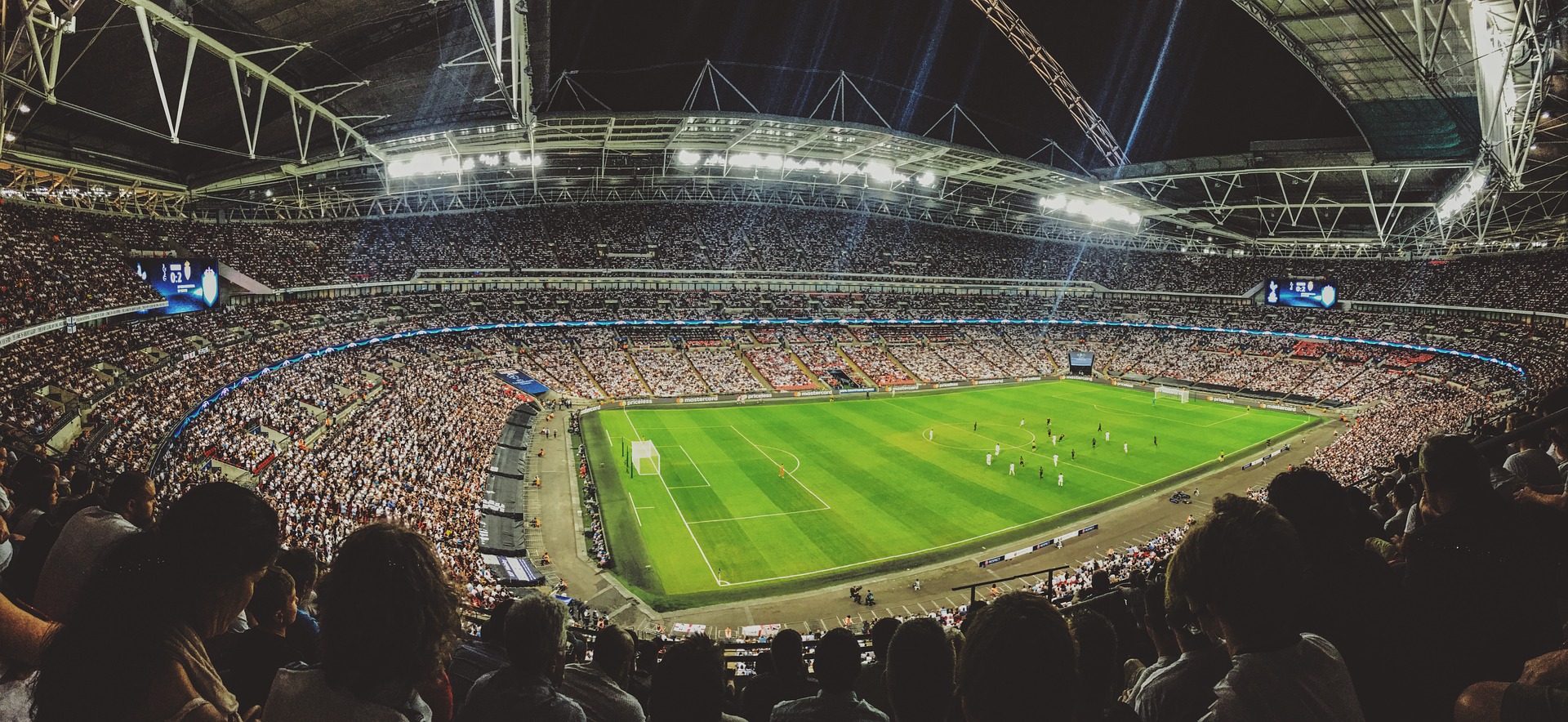Panoramic photograph of a soccer stadium. It appears that all the seats are full while players stand on the field.
