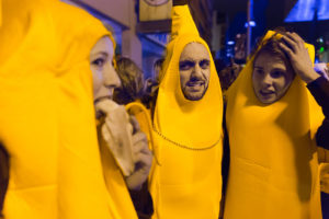 Three people shown wearing banana suits. Two look in dismay at the third, who eats a banana.