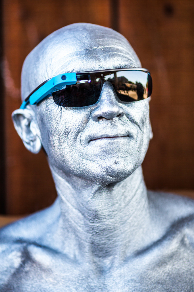 A bald man, whose entire head, face, and neck are covered in silver paint, wears a pair of Google Glass glasses and smiles.