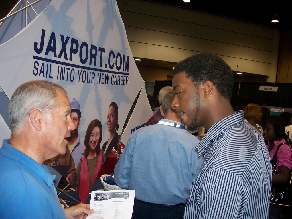 A man talks to another, younger man at a job fair. In the background is a poster advertising Jaxport.com. 
