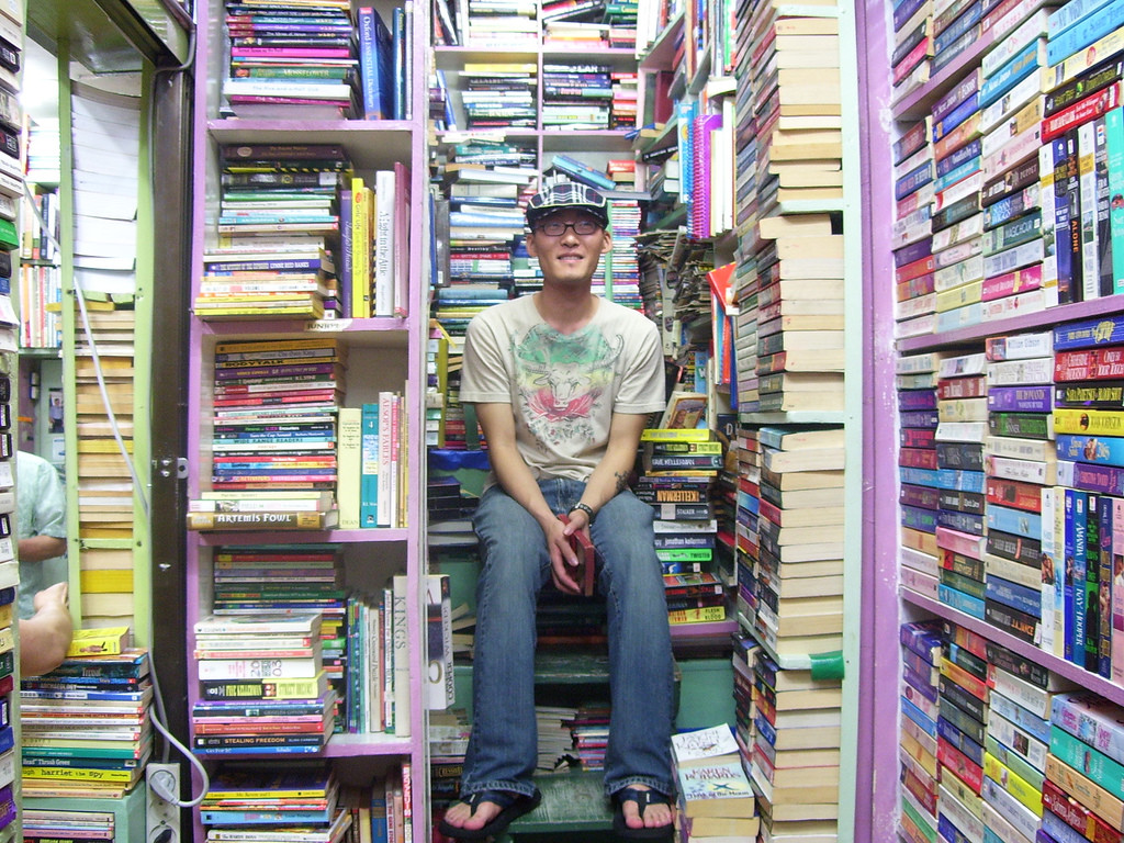 Photograph of a man sitting on a pile of books. He is surrounded by tall shelves with books haphazardly shelved. 