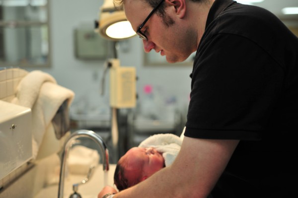 A man is shown standing in front of a sink holding a newborn baby.