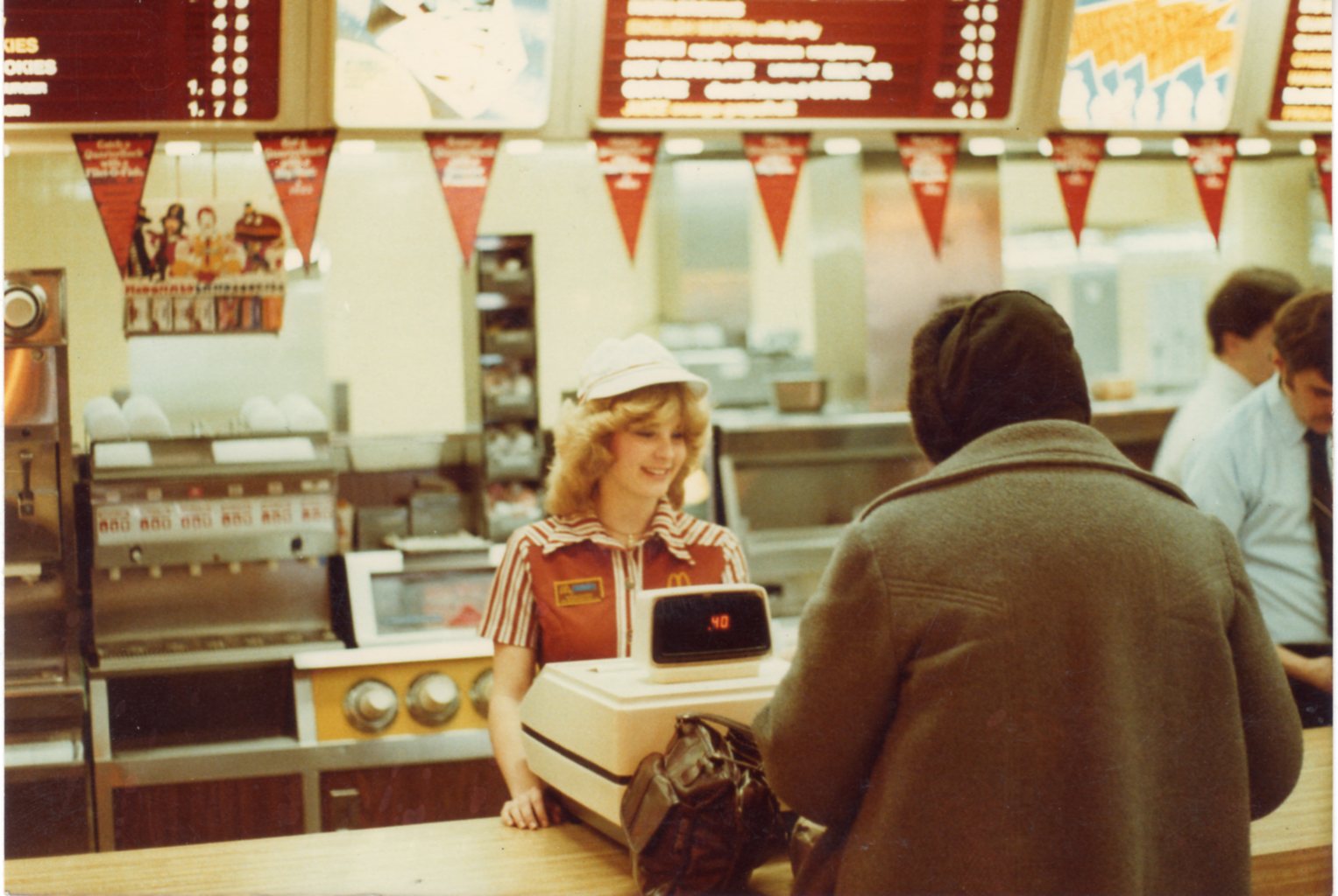 A cashier at a McDonald's is smiling and standing behind the counter and cash register taking a customer's order. 