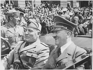 Mussolini and Hitler sitting in a car, driving past crowds of people on a street.