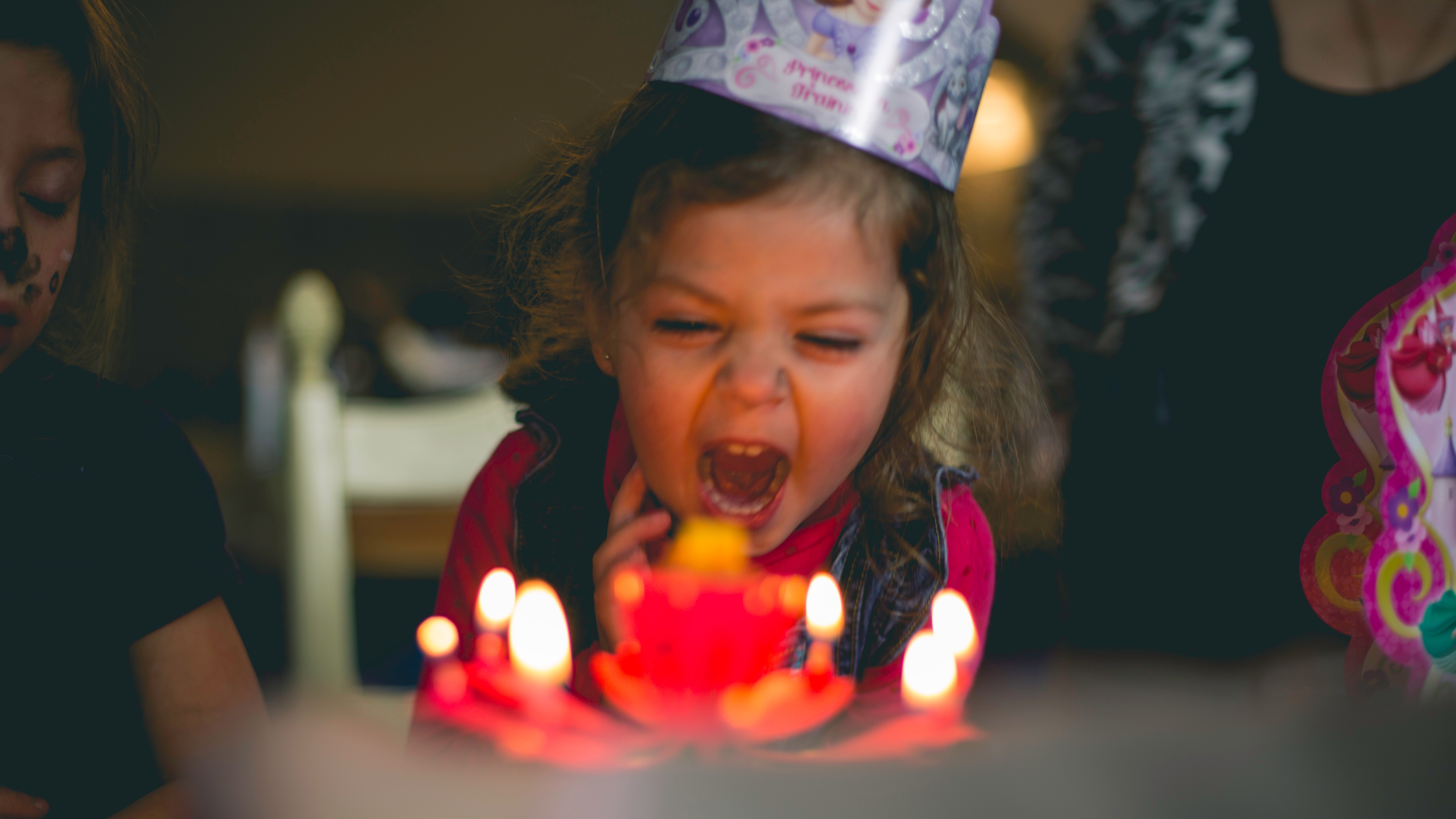 A young girl with a birthday hat is yelling in front of a cake with several lit candles. 