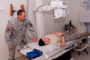 One soldier lies in an x-ray machine while another stands to the side and operates the machinery.