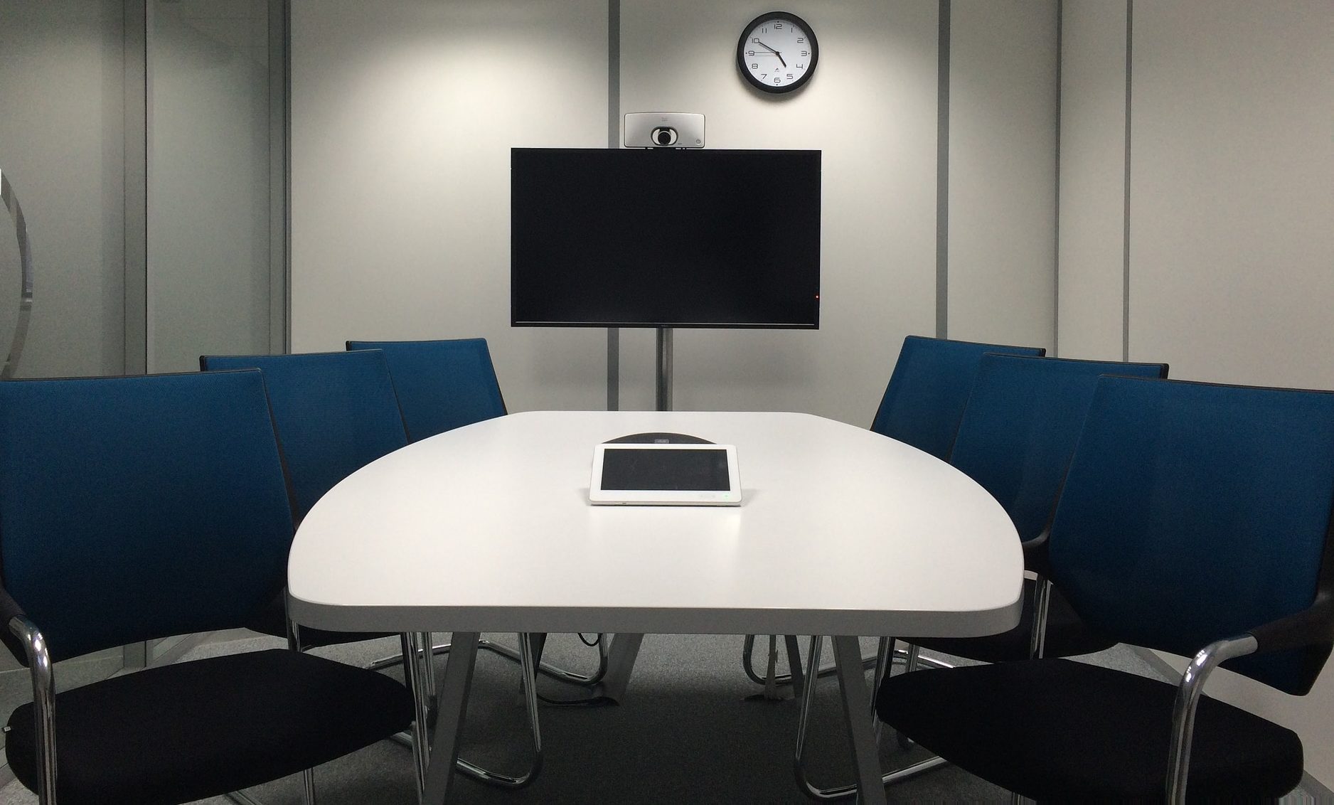 Photograph of a conference room. There is a table and several empty chairs in front of a TV. There is a microphone/speaker combination on the table.