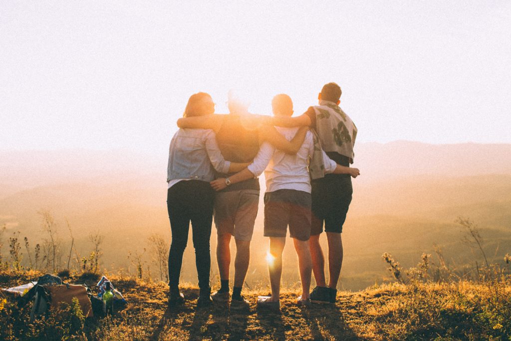A photo of four people facing away from the camera looking at the sunset. The people have their arms over one another's shoulders.