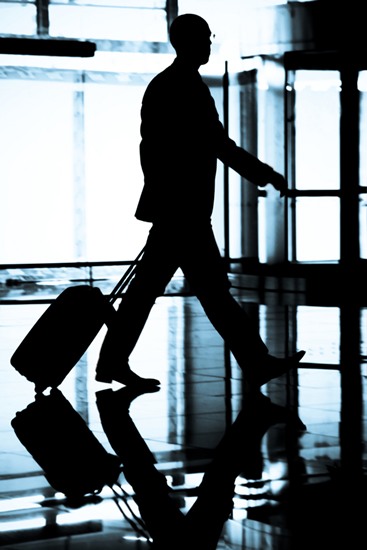 Silhouette of a man in a suit walking in an airport pulling a small suitcase.