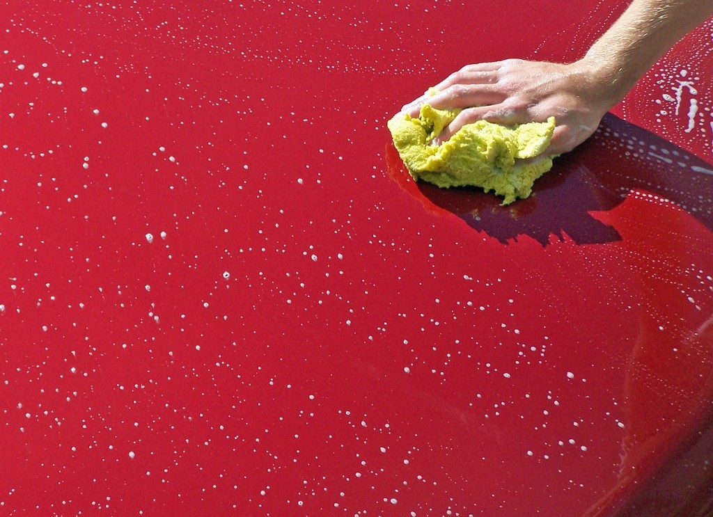 Photo showing a hand holding a sponge, washing the hood of a red car.