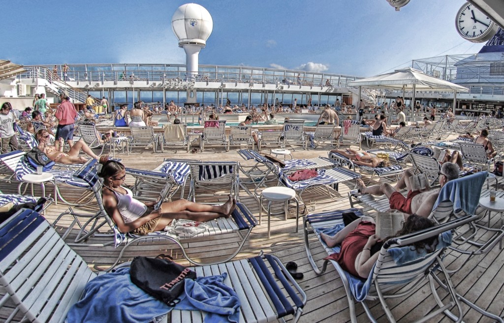 Digitally altered photo with a deck-top view of cruise ship, showing many sunbathers on lounge chairs around a large pool. The distorted colors give the picture a quiet, frozen quality. 