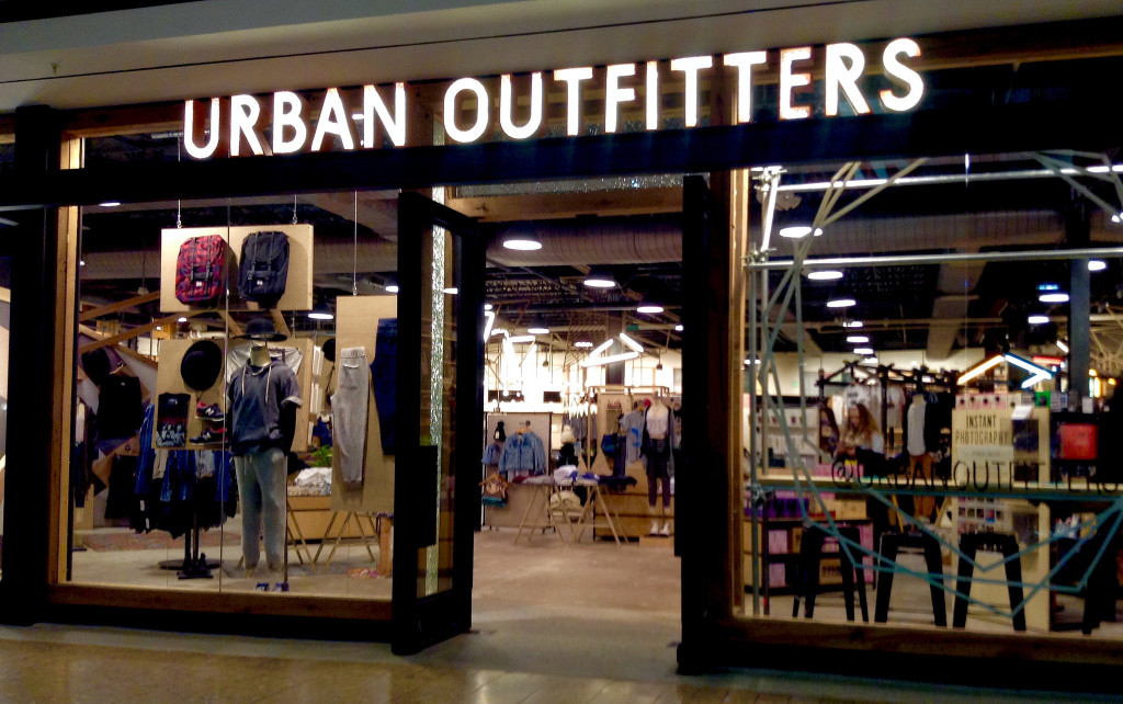 A storefront for Urban Outfitters. Near the front of the store are mannequins wearing the store's clothing. The inside of the store has industrial spotlights, a cement floor, aisles of clothing, and more mannequins.