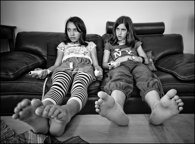 Black-and-white photo of two teenage girls lounging on couch, TV remotes in hand.