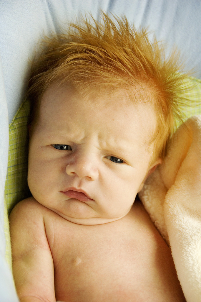 Very young infant boy with reddish orange hair scrunching is nose and frowning.