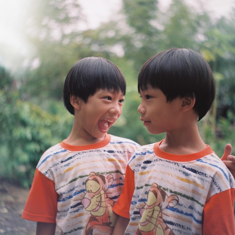 Identical twin boys look at each other, one with a straight face and the other with an open-mouth laugh.