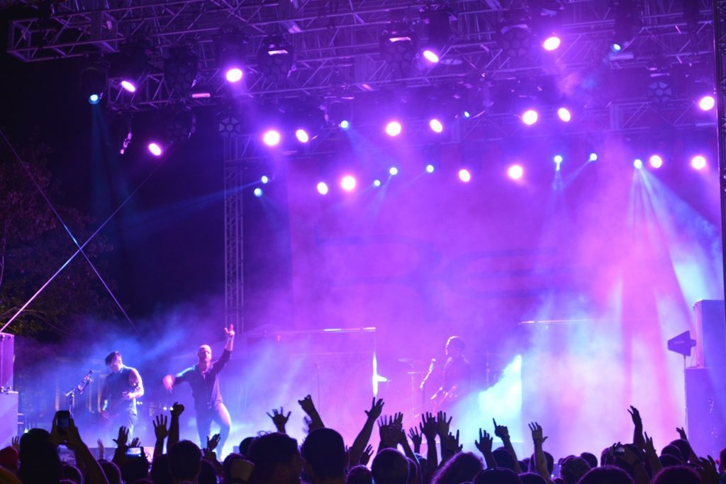 Picture from a rock concert. A band plays on the stage with blue and purple lights shining down. Fans raise their hands in the audience.