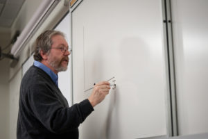 Male professor with a graying beard writing on a whiteboard, wearing a sweater and glasses.