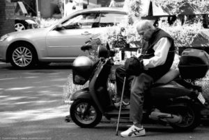 Photograph of an older man holding a cane while getting off of his moped.