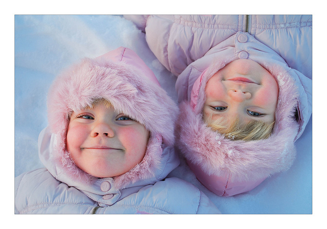 Identical twin toddler girls laying in the snow, wearing pick snowsuits.