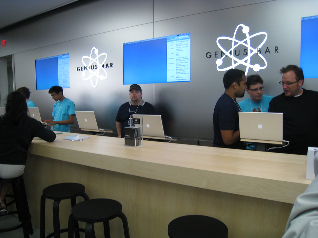 Photo of the genius bar at an apple store. Several employees sit behind a counter at Apple computers.
