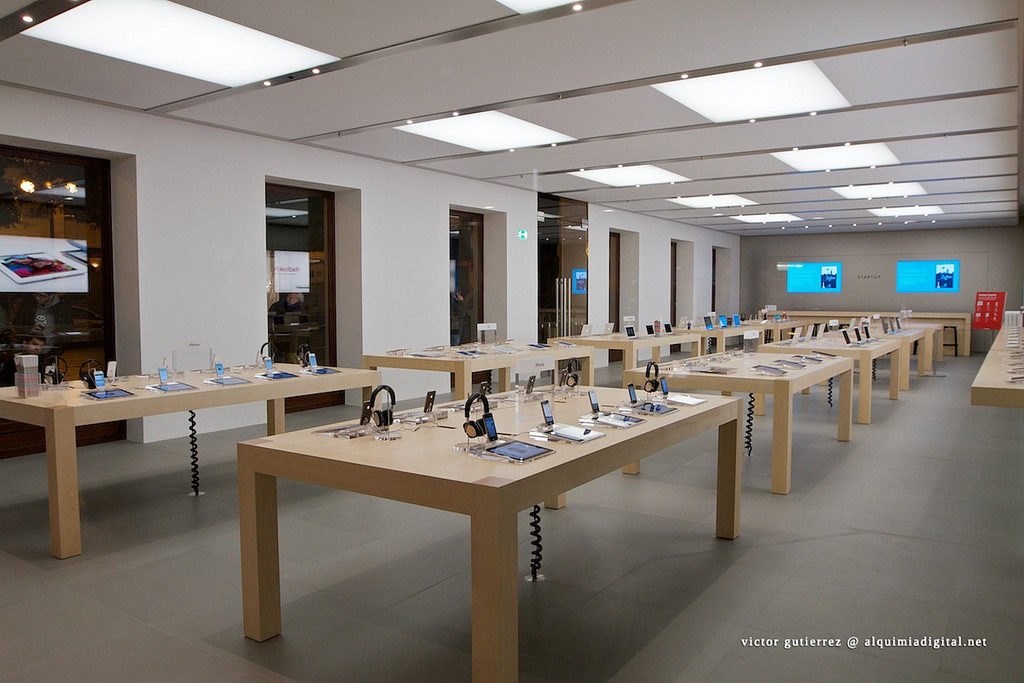 Photograph of an apple store layout. There are two rows of three tables. Each table has just a few items displayed on each surface.