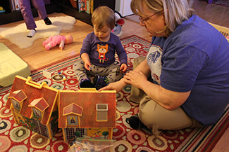 An adult and a small child are depicted sitting on a rug next to a toy house.